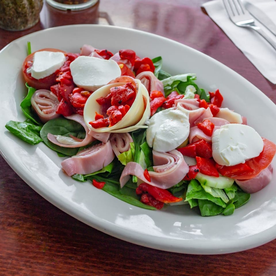 A Tuscana Pizza & Pasta salad with ham, mozzarella, roasted red peppers, and greens on a white plate.
