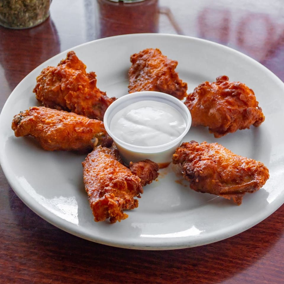 A white plate of six crispy, orange-red chicken wings surrounds a small cup of white dipping sauce.